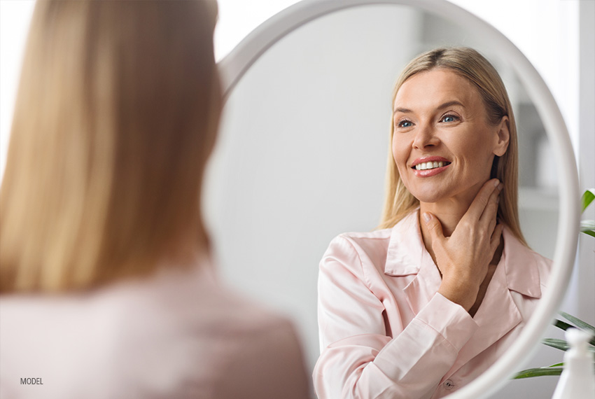 middle aged woman looking into a mirror and smiling as she touches her neck